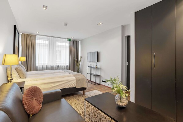 Una habitación de hotel moderna con cama, sofá, mesa de centro, televisor, armario y ventana con cortinas.