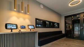 Ein Empfangsschalter mit einem Computermonitor, einem Telefon und einem digitalen Bildschirm. Eine Bank und eine Glastür führen in einen weiteren Raum.