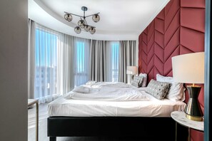Une chambre d’hôtel dotée d’un grand lit, d’un lustre moderne, d’un mur d’accent rouge et de grandes fenêtres.