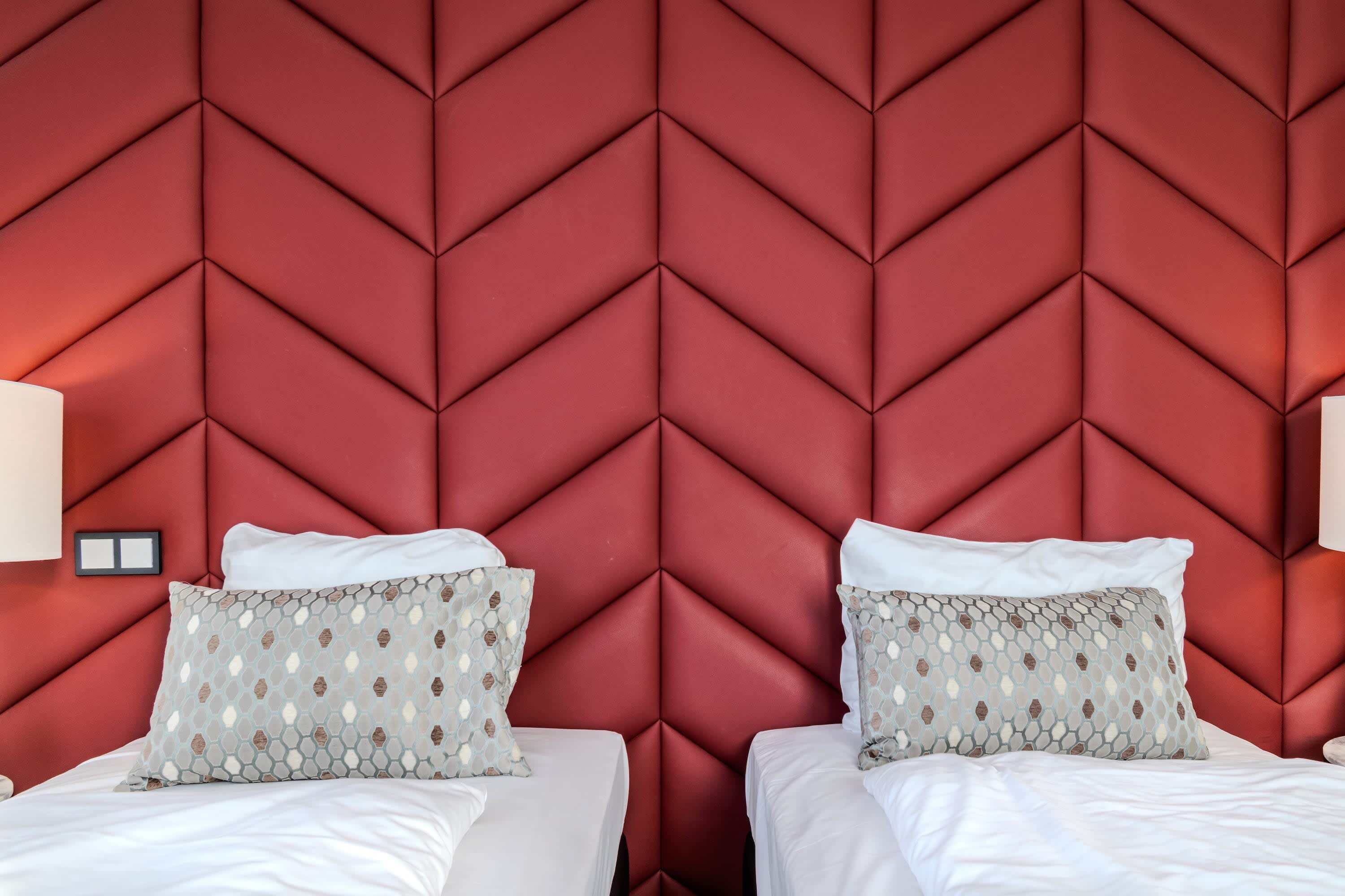 Ein Hotelzimmer mit einem roten Kopfstück mit Zickzackmuster, zwei Betten mit weißen Bettw äsche, gemusterten Kissen und Nachttischlampen.