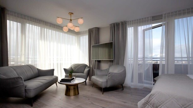 Habitación de hotel moderna con sofá, sillón y cama. Tiene un televisor en la pared y un balcón con cortinas.
