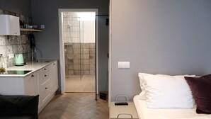 A compact hotel room with a kitchenette, a bed with white and maroon pillows, and a shower area. - Saga Studio for 2 guests in Downtown Snorri (Reykjavík)
