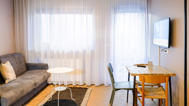 A living room with a sofa, a small round table, a wooden dining table, and a TV mounted on the wall. - Saga Studio for 2 guests in Downtown Snorri (Reykjavík)