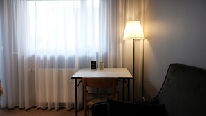 A room with a table, a chair, a lamp, and a window with sheer curtains.