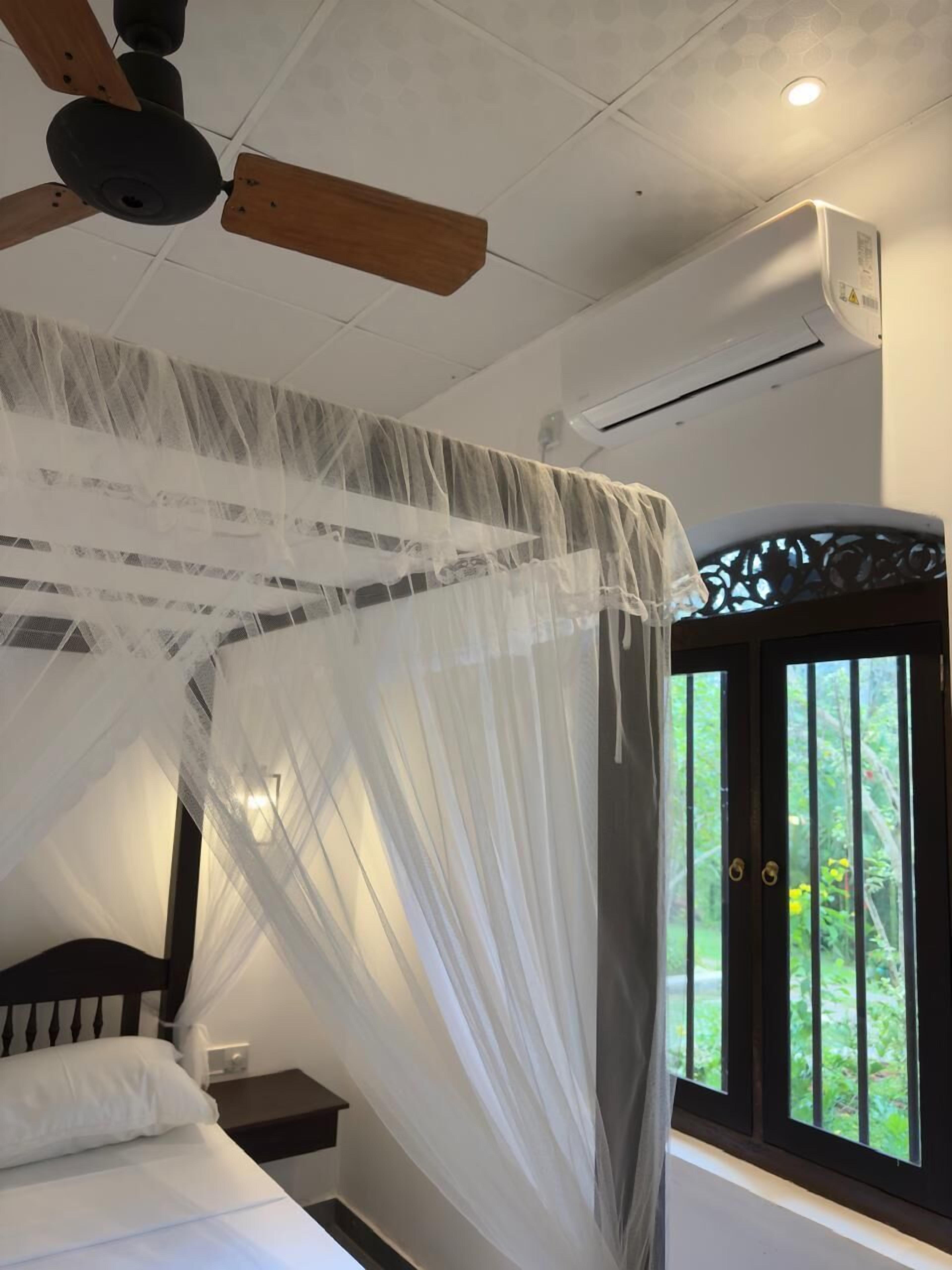 Een slaapkamer met een bed, een nachtkastje, een plafondventilator en een raam met uitzicht op groen.