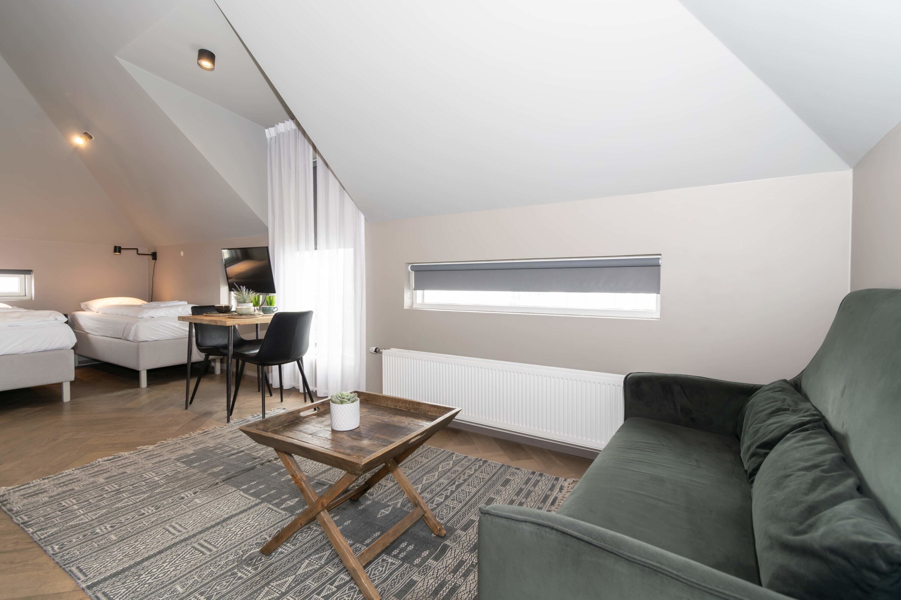 Une chambre de style loft moderne, avec un plafond en pente, un canapé vert, une table basse en bois, un coin repas avec une table et des chaises, et une grande fenêtre laissant entrer la lumière naturelle.