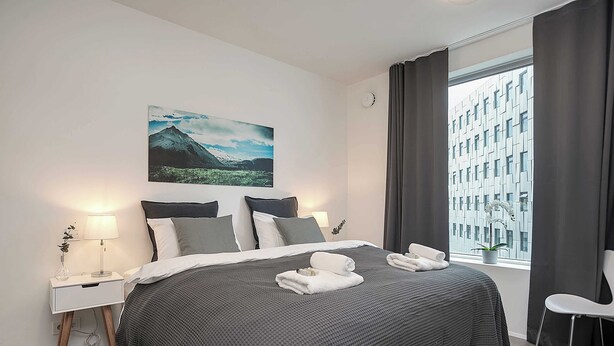 A modern hotel room with a large bed, a nightstand with a lamp, a window with curtains, and a cityscape view.