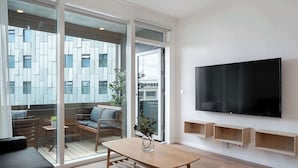 A modern living room with a wooden table, a sofa, a TV mounted on the wall, and a sliding glass door leading to an outdoor area with a view of a building.
