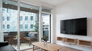Una sala moderna con una mesa de madera, un sofá, un televisor montado en la pared y un ventanal corredero que da a un área exterior con vistas a un edificio.