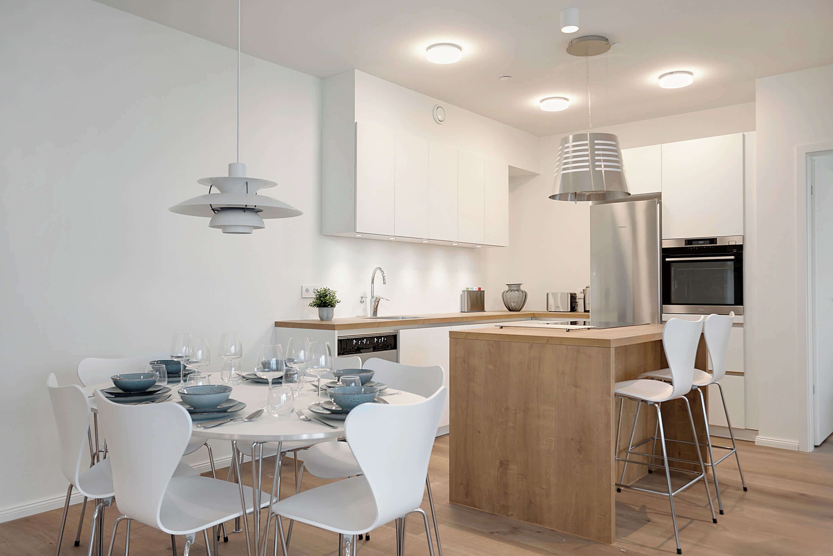 Une cuisine moderne avec des meubles blancs, un îlot central en bois et un coin repas comprenant des chaises blanches et des bols bleus.