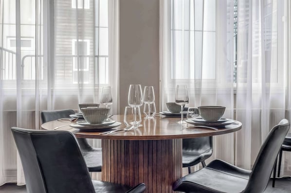 A round dining table with chairs, set for a meal, in a room with large windows and sheer curtains.