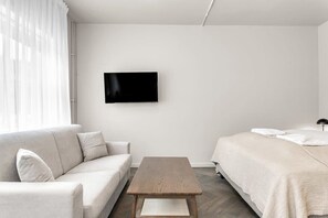 A modern, minimalist hotel room with a sofa, a wooden table, a bed with a white duvet, and a flat-screen TV mounted on the wall.