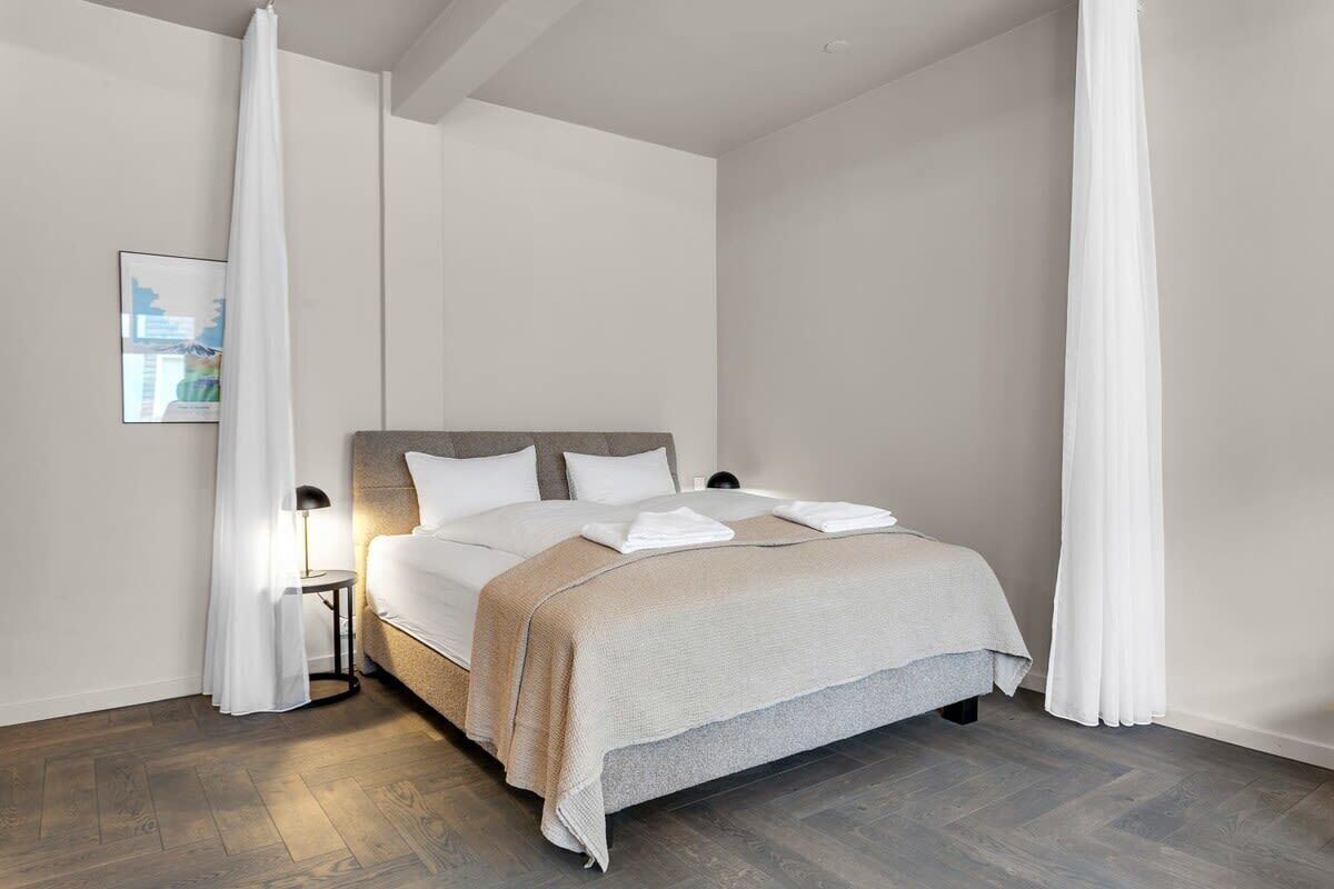 A modern hotel room with a large bed, white bedding, and a small bedside table with a lamp.