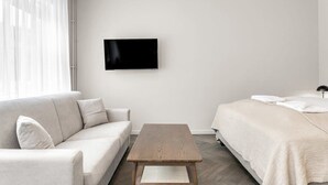 Quarto de hotel moderno e minimalista, com sofá, mesa de madeira, cama com edredão branco e televisão de tela plana fixada na parede.
