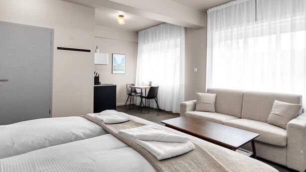 A modern hotel room with two beds, a sofa, a small table, a desk, and a dining area.