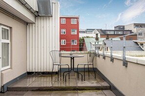 A small outdoor patio with a table and two chairs, a metal railing, and a view of a red building in the background.