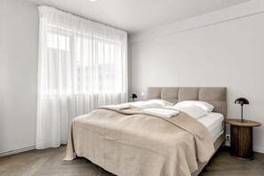 Un dormitorio con cama, mesita de noche, lámpara y ventana con cortinas translúcidas.