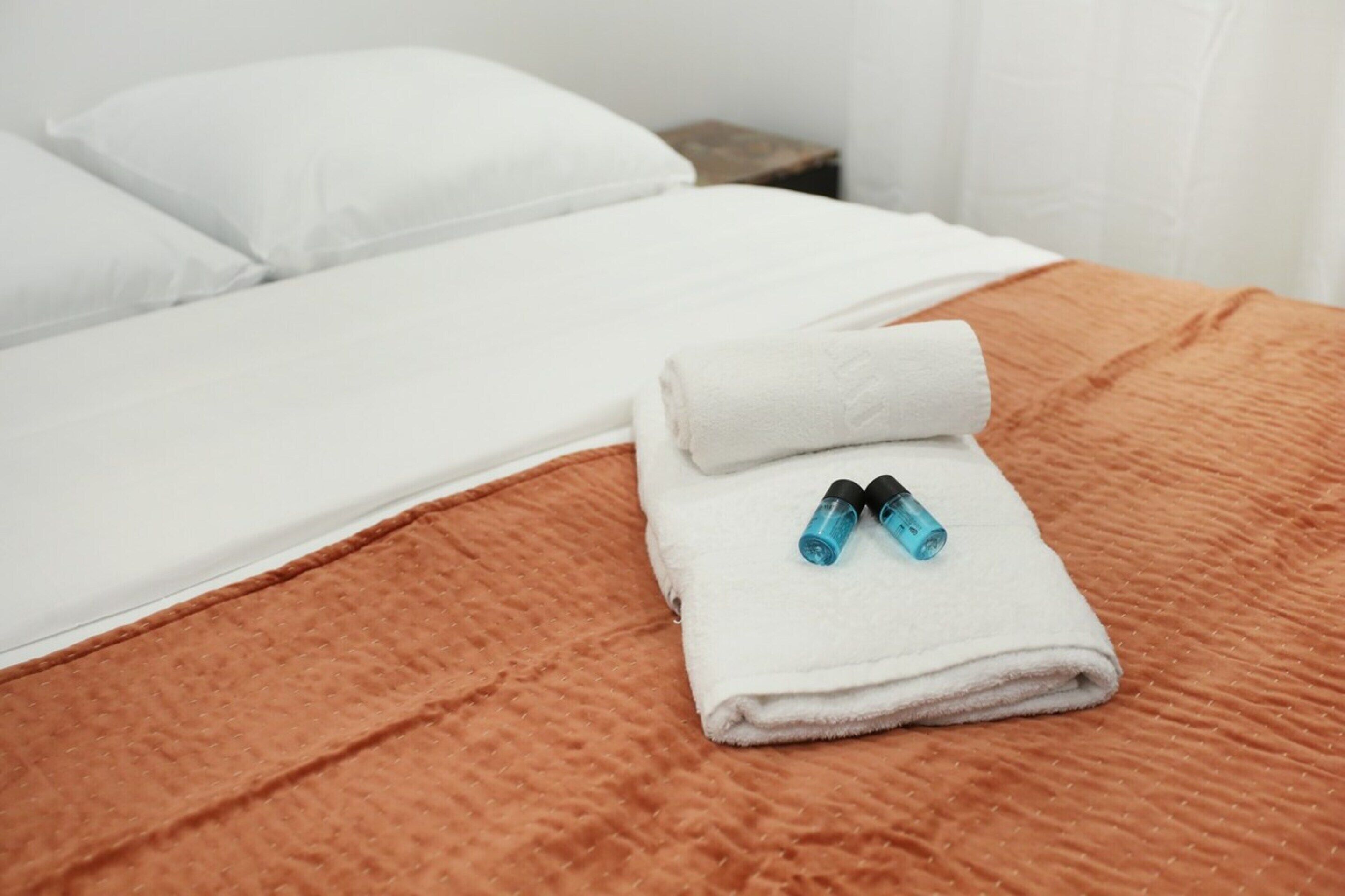Cama bem arrumada com uma toalha branca dobrada e dois frascos azuis.