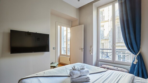A bedroom with a bed, a TV mounted on the wall, a window with curtains, and a view of buildings outside.