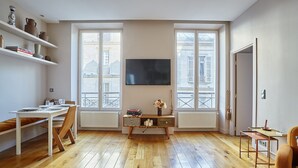A modern living room with a wooden floor, a flat-screen TV mounted on the wall, a dining table with chairs, and a small side table with a vase of flowers.