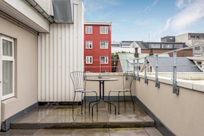 Une petite terrasse extérieure avec une table et deux chaises, une rambarde en métal et, en arrière-plan, une vue sur un bâtiment rouge.