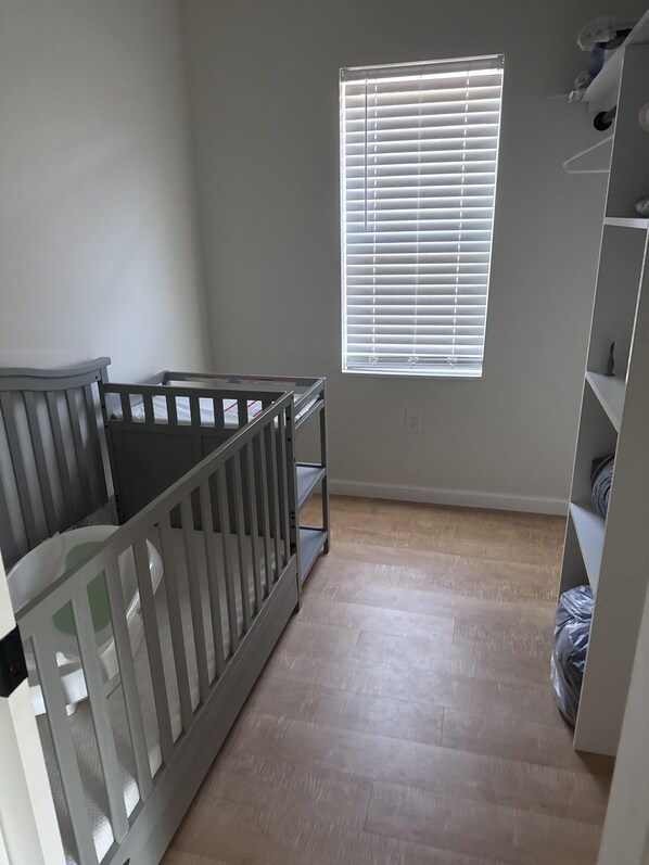 A room with a crib, a window with blinds, and a shelving unit.