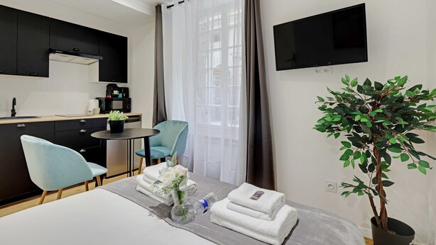 A hotel room with a bed, a small dining table, a chair, a TV, a potted plant, and a view of the outside through a window.