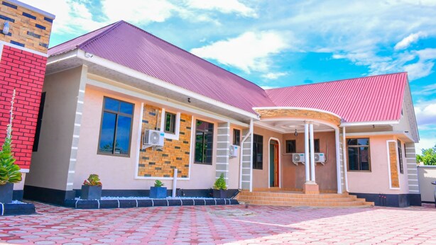 A two-story building with a red roof, a brick facade, and a covered entrance with steps. - DOM PRECIOUS LODGE (Dodoma)