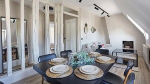 Un comedor con una mesa negra dispuesta para cuatro personas, rodeada de sillas. Hay dos espejos en la pared, un sofá y una pequeña mesa con un televisor.