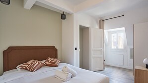 Una cama bien hecha con cabecera de madera, ropa de cama blanca y un par de toallas dobladas. A la derecha, una mesita de noche con una lámpara. La habitación tiene paredes blancas, piso de madera y una puerta que da a otra habitación.