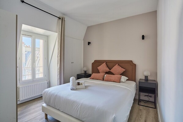 A bedroom with a large bed, a nightstand, a window with curtains, and a radiator. - Sublim Apartment Beaubourg/le Marais (Paris)
