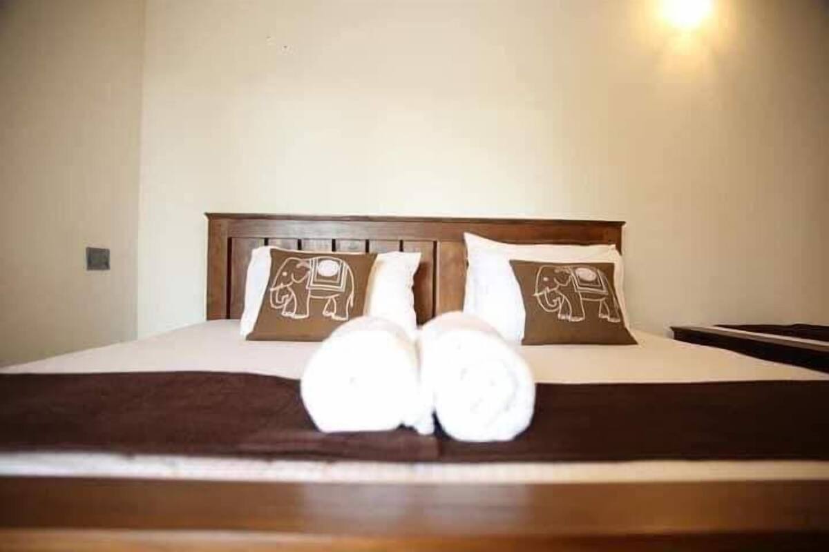 A neatly made bed with two pillows featuring elephant designs, a brown blanket, and a pair of rolled towels on the bed.