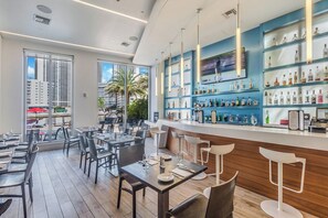 A modern restaurant with a bar area, large windows, and a view of palm trees. - Waterfront Pool • Beach Service • 7th Floor (Hallandale Beach)
