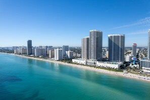 A coastal cityscape with high-rise buildings, a beach, and a clear blue sky. - Waterfront Pool • Beach Service • 7th Floor (Hallandale Beach)