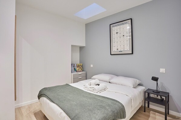 Un dormitorio moderno con tragaluces, un cartel enmarcados en la pared, una mesita de noche con lámpara y toallas ordenadas sobre la cama.
