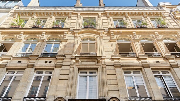 Un immeuble à plusieurs étages, de style architectural classique, caractérisé par des fenêtres en arc de cercle et des balcons décoratifs ornés de plantes en pot.