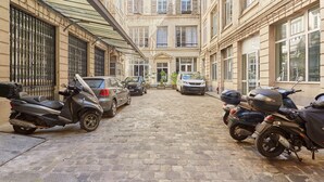 Une ruelle étroite pavée, bordée de bâtiments aux grandes fenêtres et aux volets, où sont garées des scooters et des voitures.