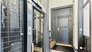 Un couloir étroit, avec un portail en métal, une porte en bois et une petite marche menant à une autre porte surmontée d’un paillasson.
