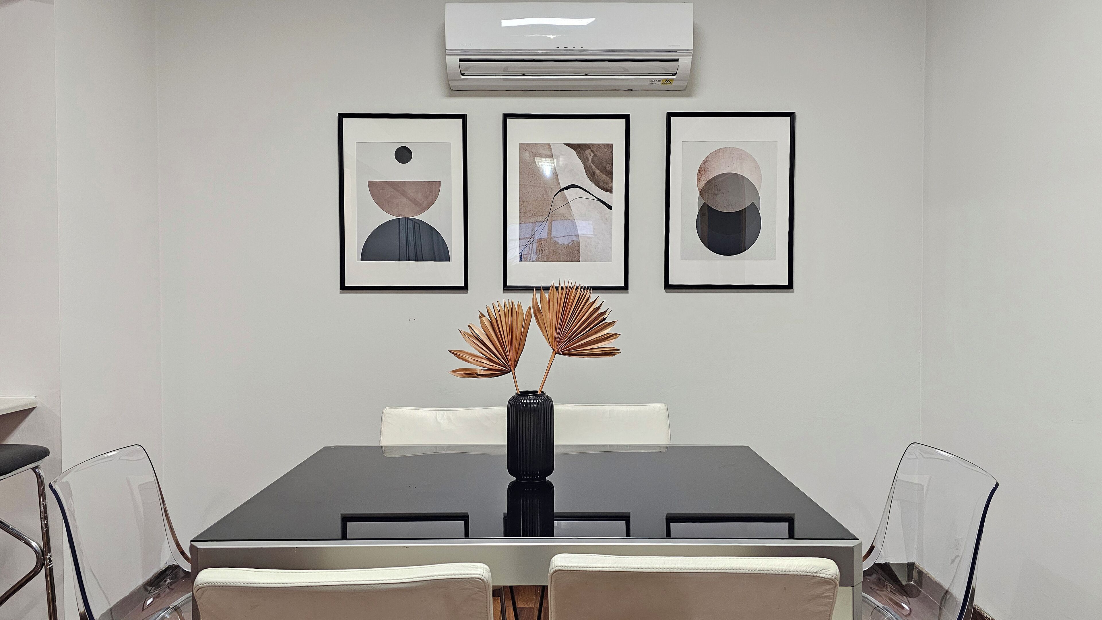 A dining area with a black table, white chairs, and a vase with decorative leaves. There are three framed artworks on the wall above the table.