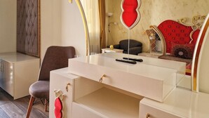 A bedroom with a white vanity, a brown chair, a red heart-shaped mirror, and a red tufted headboard.