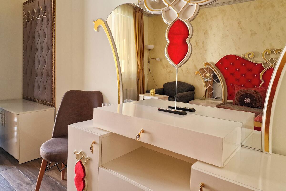 Un dormitorio con un tocador blanco, una silla marrón, un espejo con forma de corazón rojo y un cabecero tapizado en rojo.