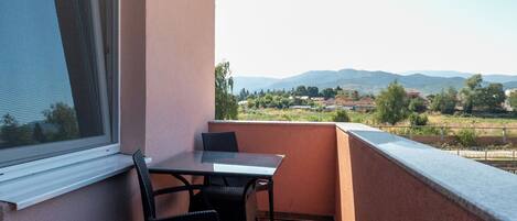 Un balcón con mesa y sillas, una ventana y vistas a un pueblo y montañas.