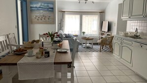 A dining area with a table set for breakfast, a kitchen with white cabinets, and a living area with a sofa and a painting of birds on the wall. - Ferienwohnung in St. Peter-ording (St. Peter-Ording)
