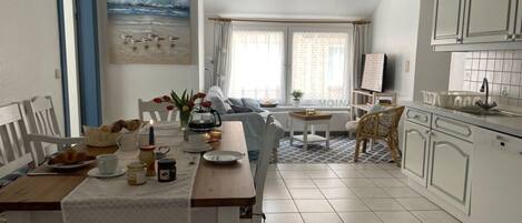 A dining area with a table set for breakfast, a kitchen with white cabinets, and a living area with a sofa and a painting of birds on the wall.