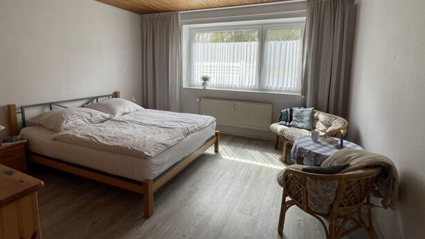 A bedroom with a bed, a chair, a small table, a window with curtains, and a ceiling fan. - Ferienwohnung in St. Peter-ording (St. Peter-Ording)