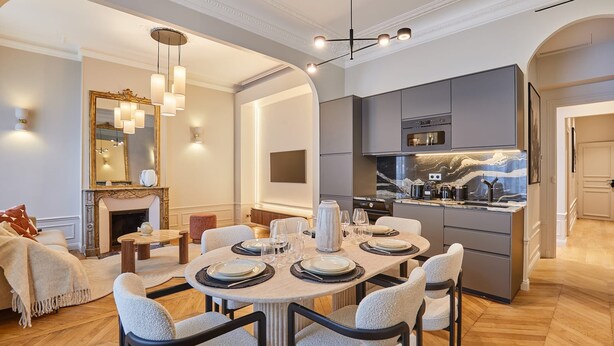 Un comedor moderno con una mesa redonda dispuesta para seis personas, una cocina con armarios grises y una chimenea con un espejo encima.