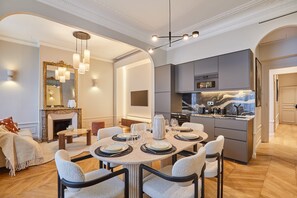 Uma sala de jantar moderna com uma mesa redonda posta para seis pessoas, uma cozinha com armários cinzentos e uma lareira com um espelho em cima.