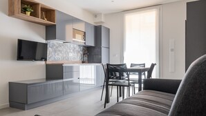 Una sala moderna con un sofá gris, una mesa de comedor con sillas, una zona de cocina con armarios grises y un televisor de pantalla plana montado en la pared.