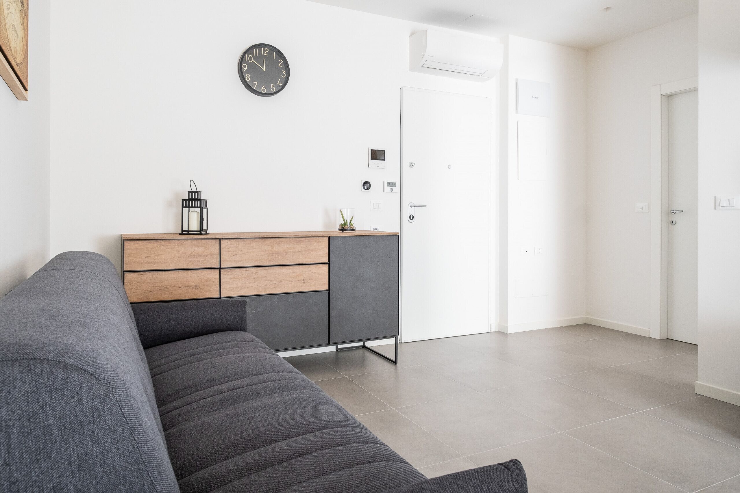 Una sala de estar moderna con un sofá gris, una mesita de madera y un mueble empotrado. Un reloj de pared y un termostato están montados en la pared.