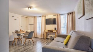 Un salon moderne avec un canapé gris, une table à manger avec des chaises, un petit bureau et un téléviseur fixé au mur.